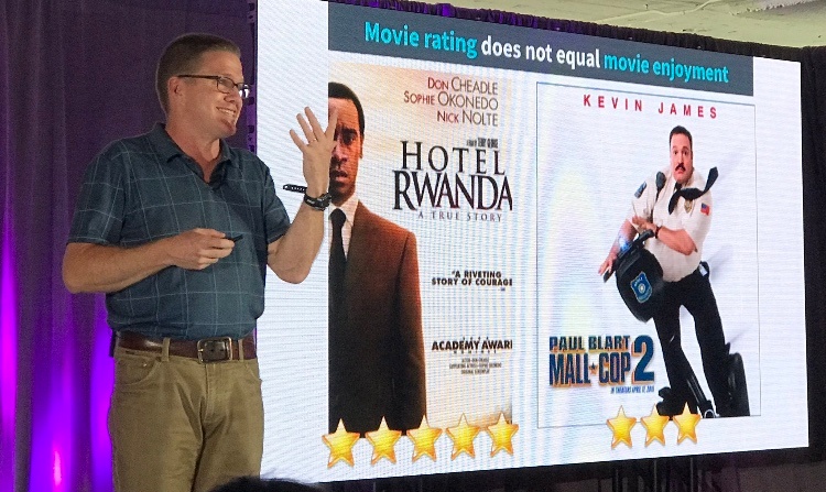 A smiling man wearing glasses and a polo shirt stood beside a screen presenting a comparison between two different film posters