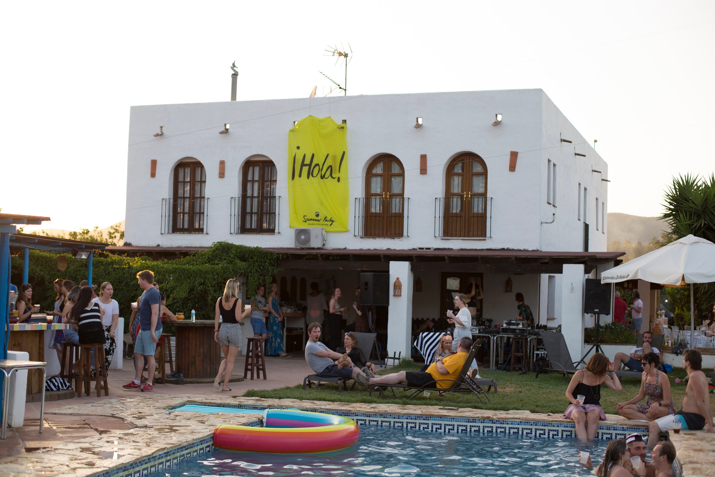 A photo outside of the hotel/villa with some people in the pool and others stood having a chat by the bar.