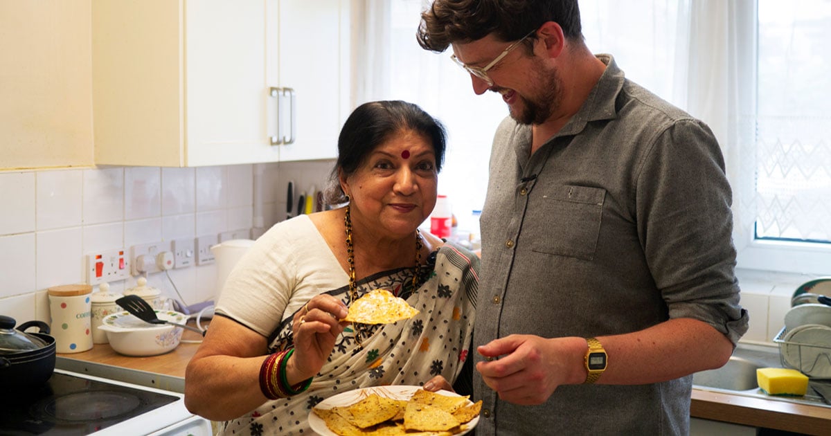A young man and an older woman stand smiling in a kitchen sharing a plate of flat breads.