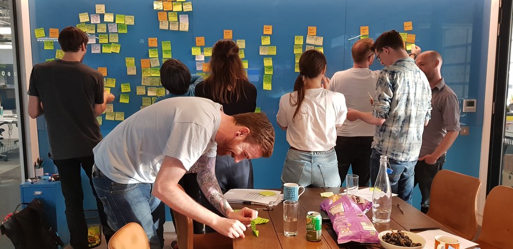 A photo of a meeting as everyone looks at the post it notes stuck up while one person rushes to finish his note.